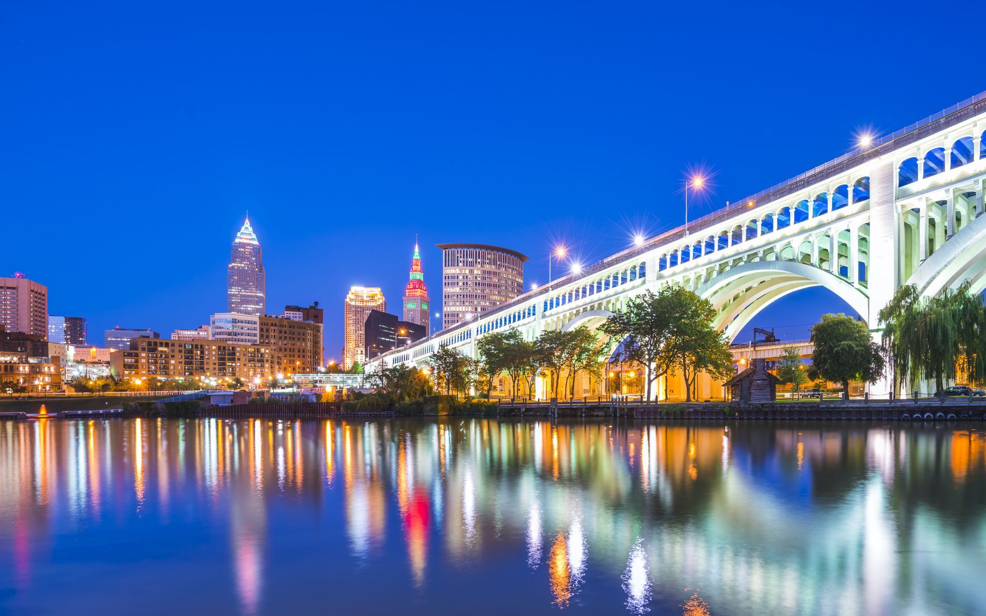 City skyline at dusk with a lit bridge reflecting in calm water.