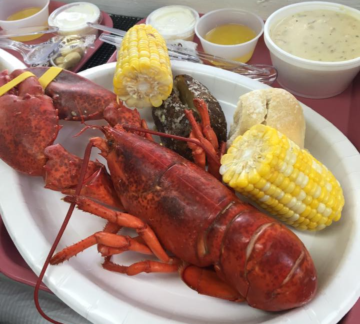 Cooked lobster, corn, potato, bread, and soup served on a white plate.