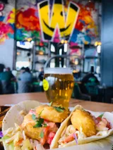 Two tacos with fried fish, beer glass, colorful mural background.