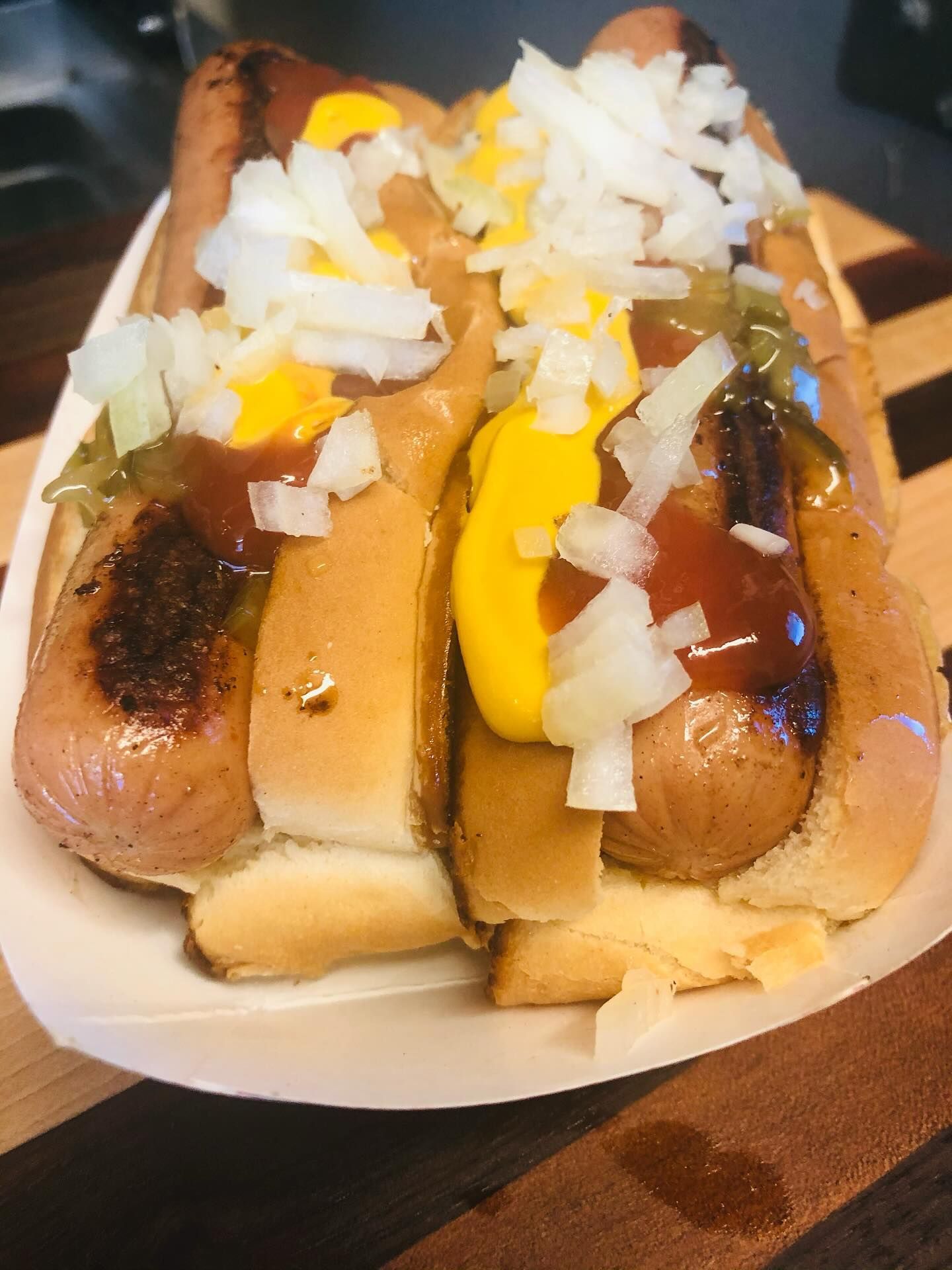 Two grilled hot dogs in buns, topped with mustard, ketchup, onions, and relish, in a paper tray.
