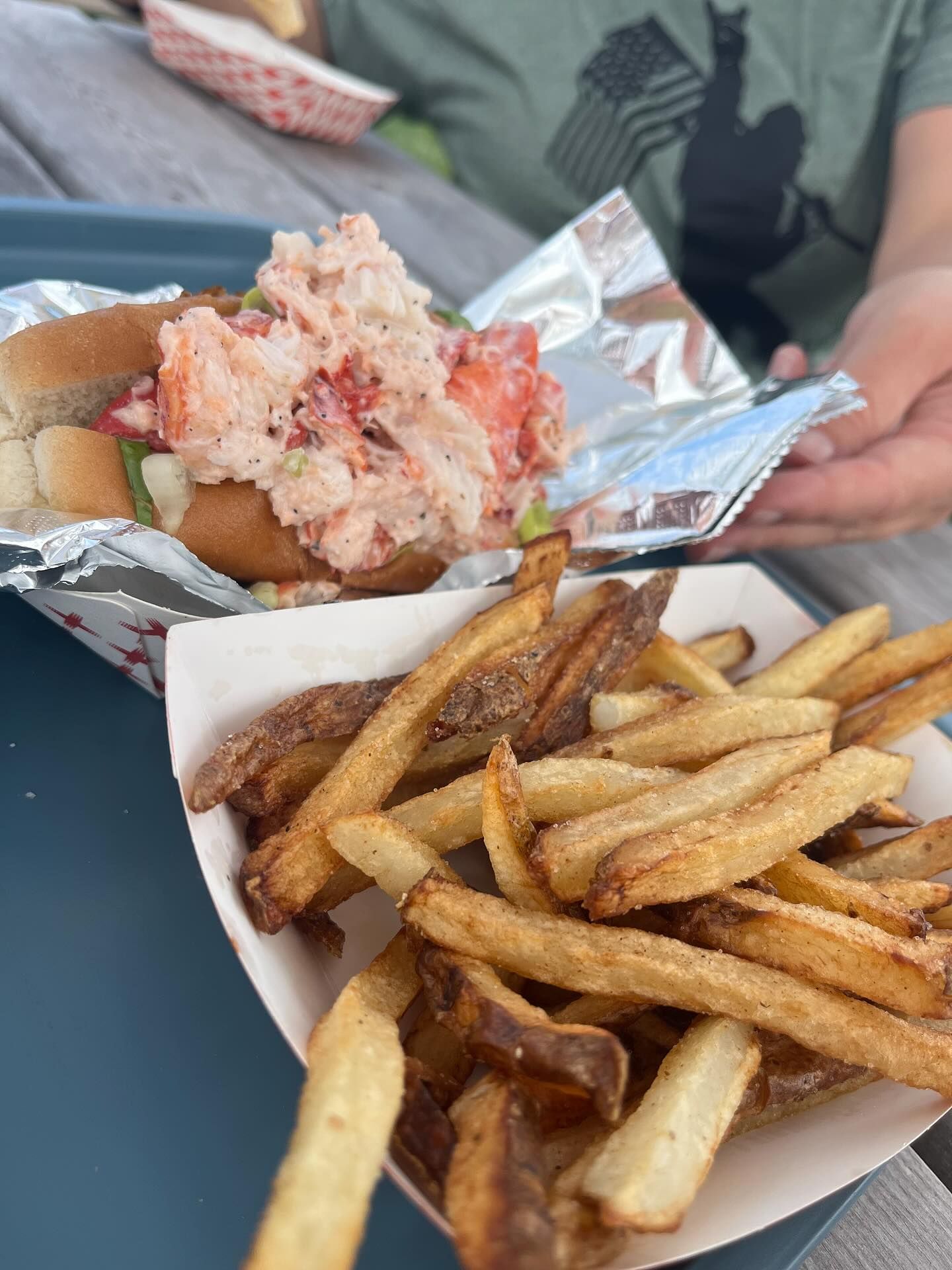 Lobster roll with fries in a paper boat on a tray. A person holds the foil-wrapped sandwich. Outdoor setting.