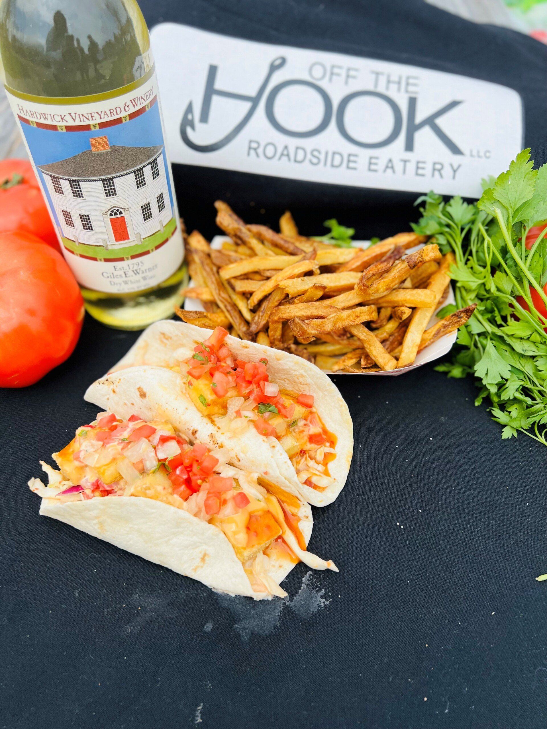 Fish tacos with fries, Off The Hook roadside eatery branding, white wine, tomatoes, and cilantro on a black surface.