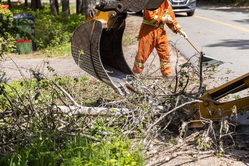 A worker in orange coveralls uses a front-end loader to clear debris from a roadside.
