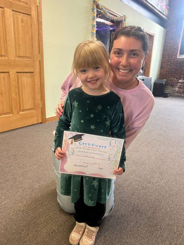 Smiling child holding a certificate while a woman kneels behind her in a carpeted room.