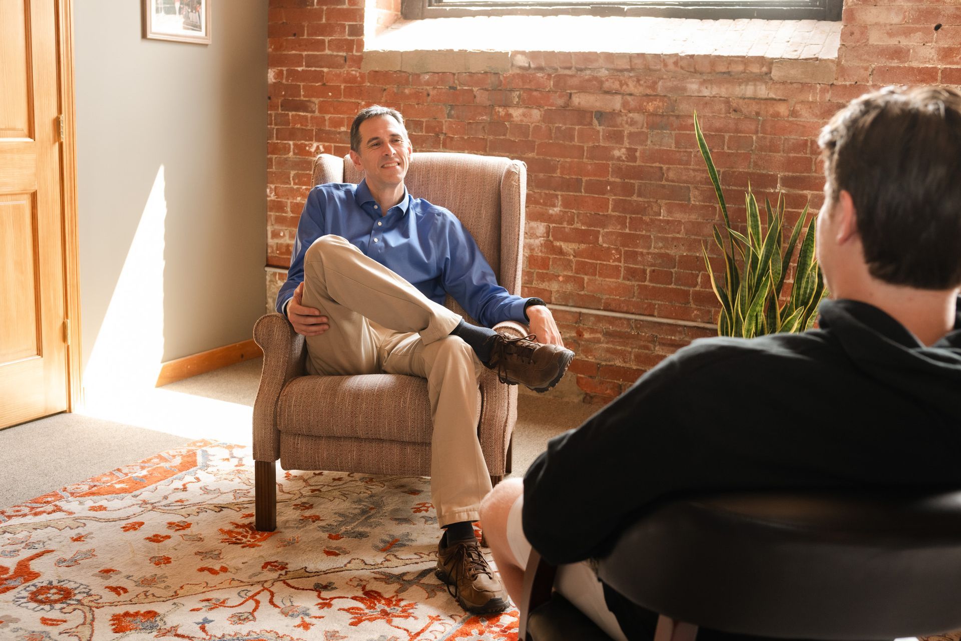 Two people sit in chairs facing each other in an office with exposed brick walls, engaged in a conversation.