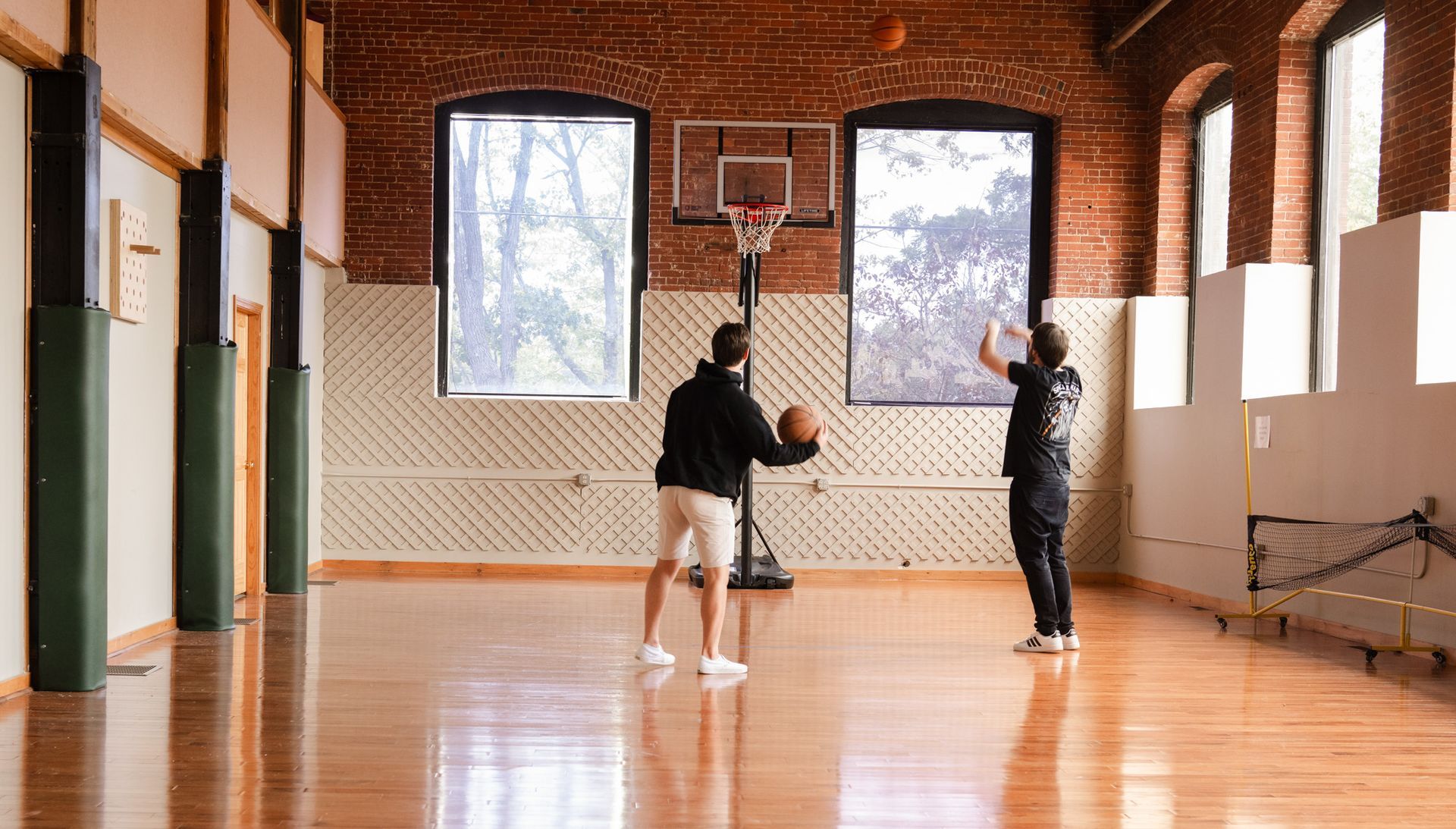 Two people playing basketball in an indoor gym with brick walls and large windows.