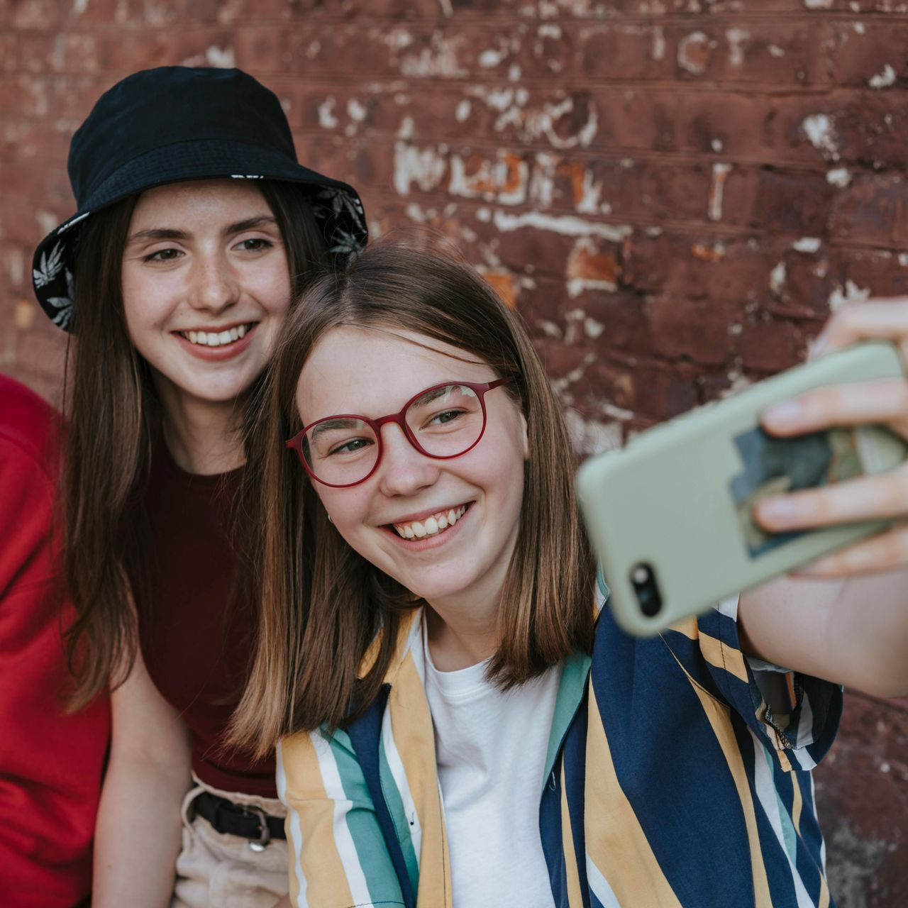 Two smiling young women take a selfie in front of a brick wall. One wears glasses and striped shirt.