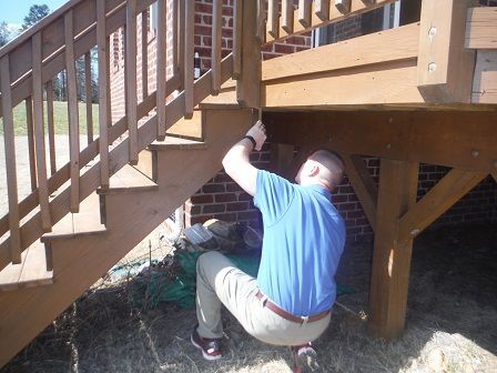 Man working on deck stairs, reaching up to adjust a piece of wood. Brown and beige tones.