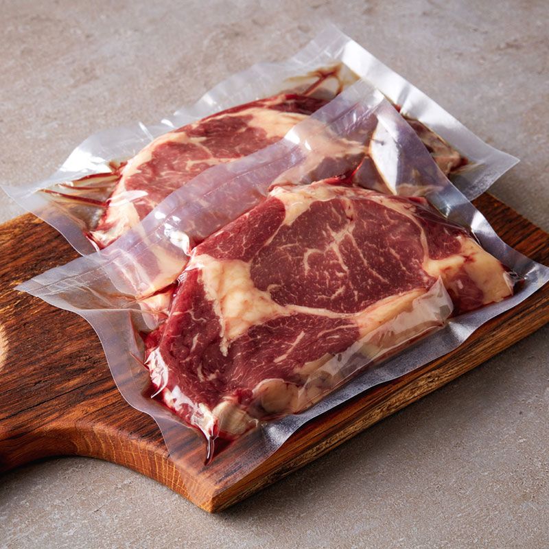 Two steaks wrapped in plastic on a wooden cutting board.