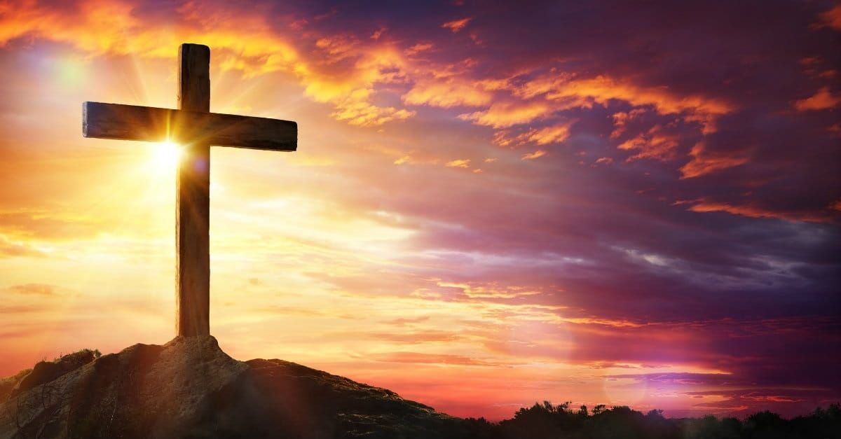 A cross on top of a hill at sunset with the sun shining through the clouds.