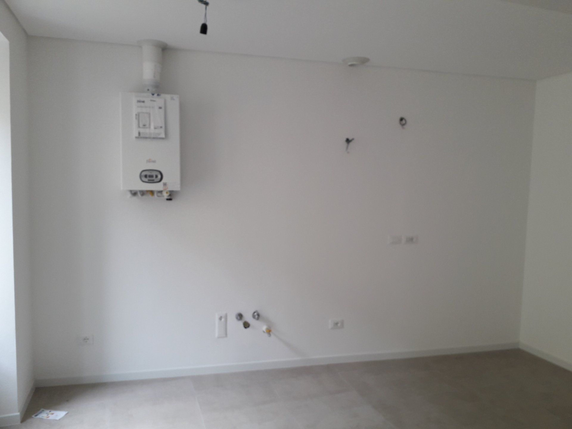 Stanza bianca vuota con caldaia a muro, prese elettriche e pavimento nudo in un interno minimalista.