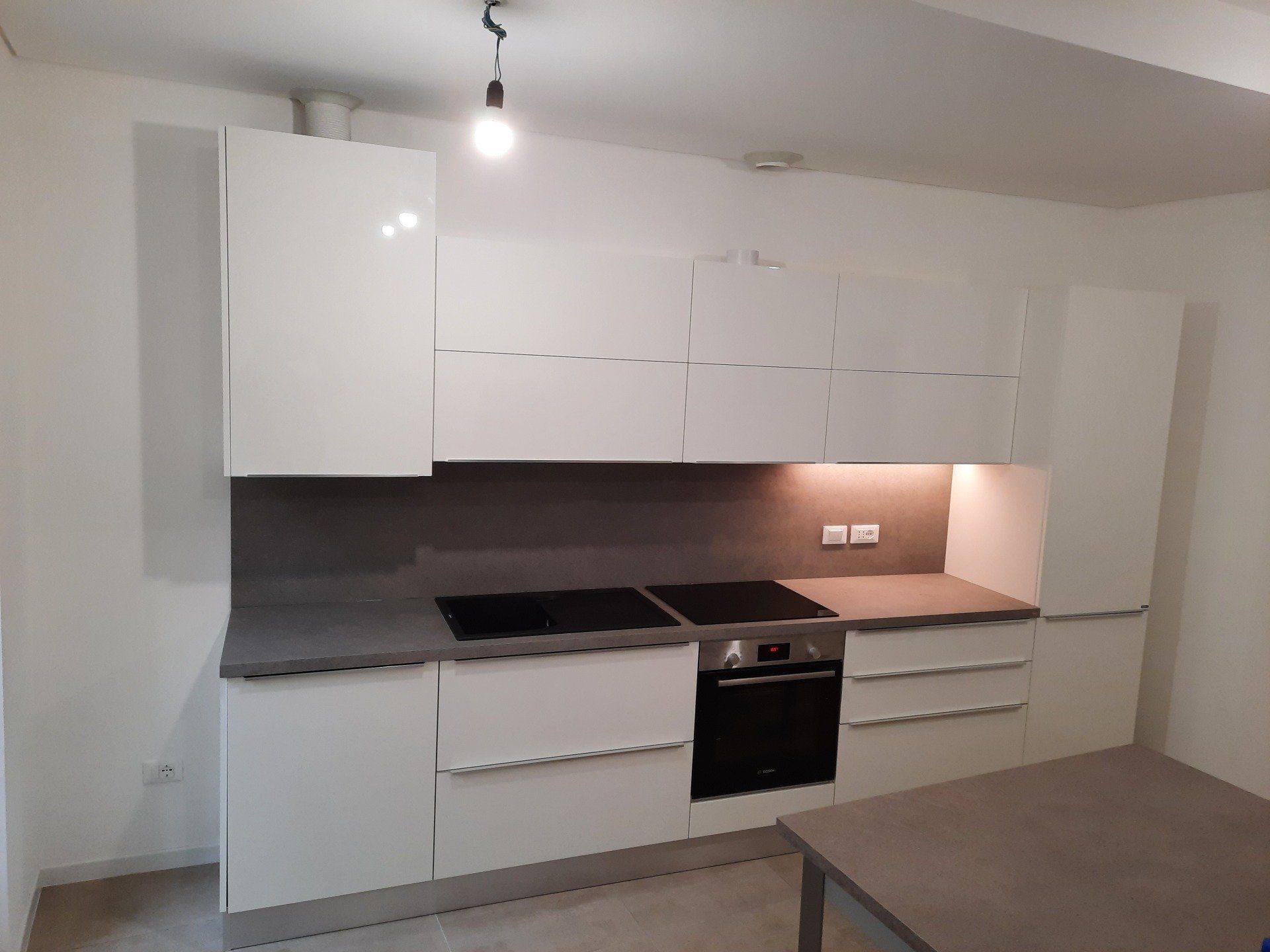 Cucina moderna bianca con piano di lavoro nero, forno a incasso e illuminazione sottopensile.