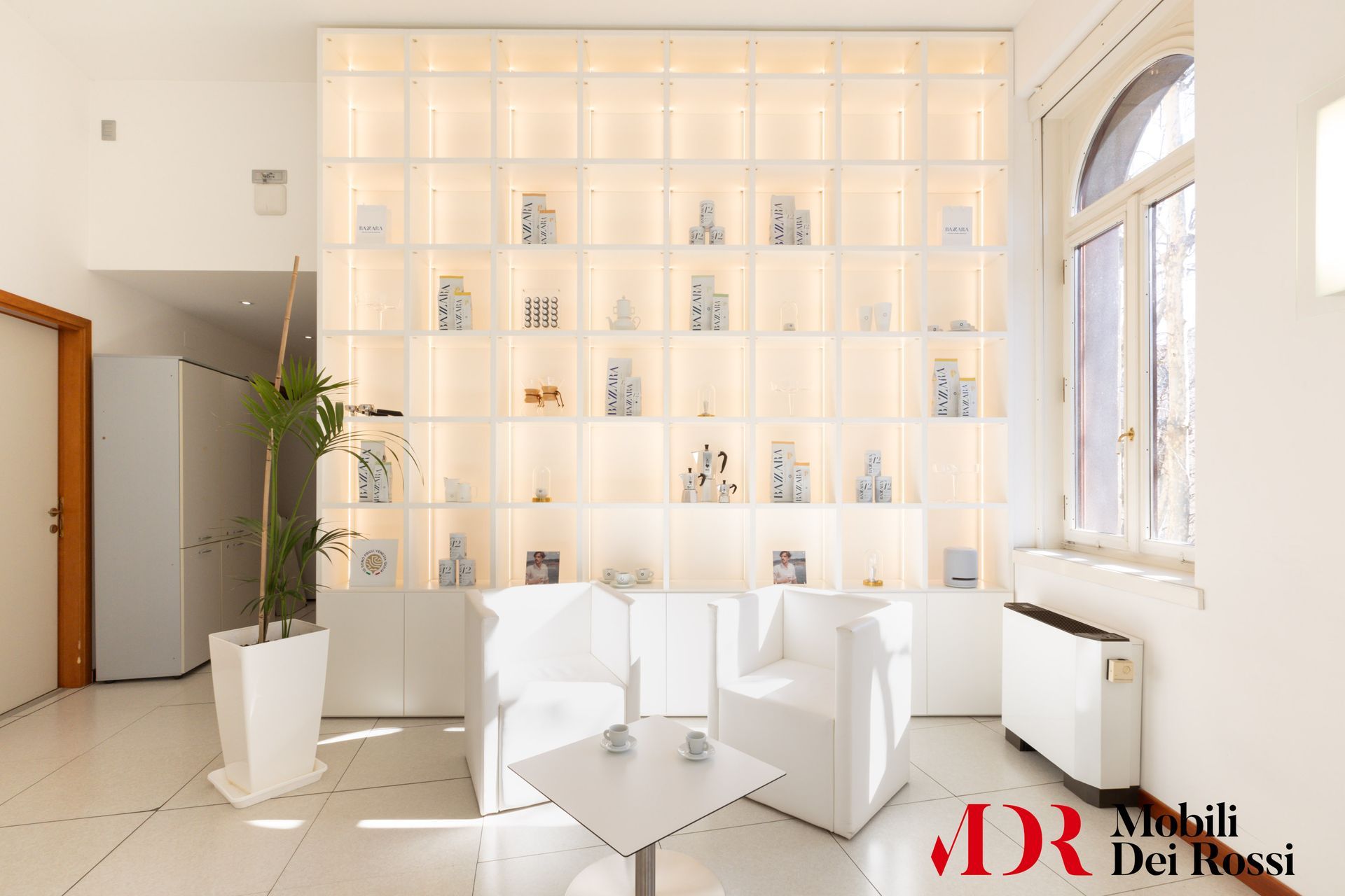 Luminoso showroom moderno di colore bianco con scaffali, sedie e una pianta alta vicino a una finestra.