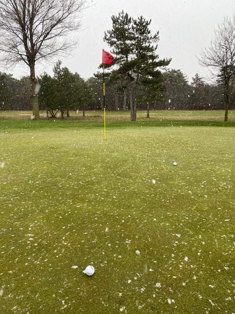Campo da golf innevato con bandierina gialla e segnaposto rosso sotto un cielo nuvoloso.