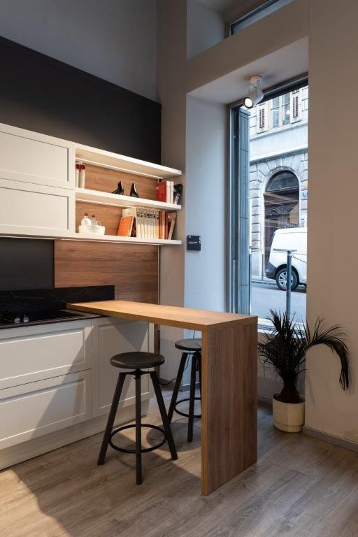 Angolo cucina moderno con mobili bianchi, mensole in legno e due sgabelli accanto a un bancone con finestra.