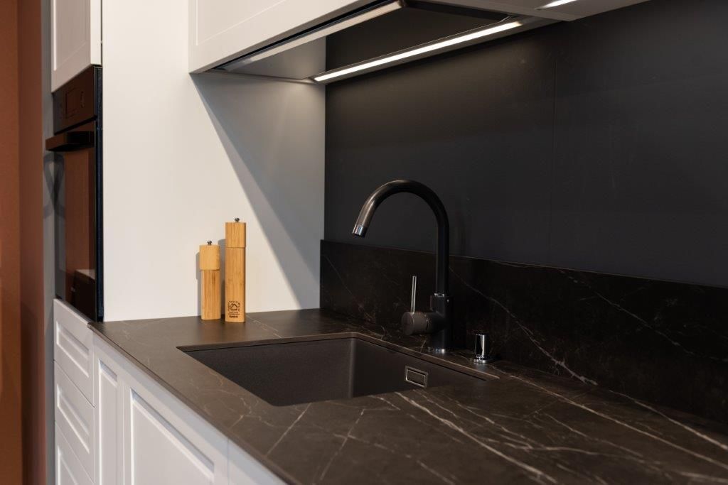 Cucina moderna con piano di lavoro nero, lavello, rubinetto opaco, illuminazione sottopensile e mobili in legno chiaro.