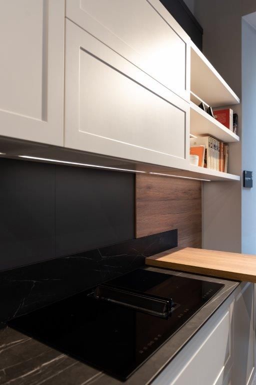 Cucina moderna con piano cottura nero, mobili bianchi, paraschizzi in legno e una piccola mensola in legno sul piano di lavoro.