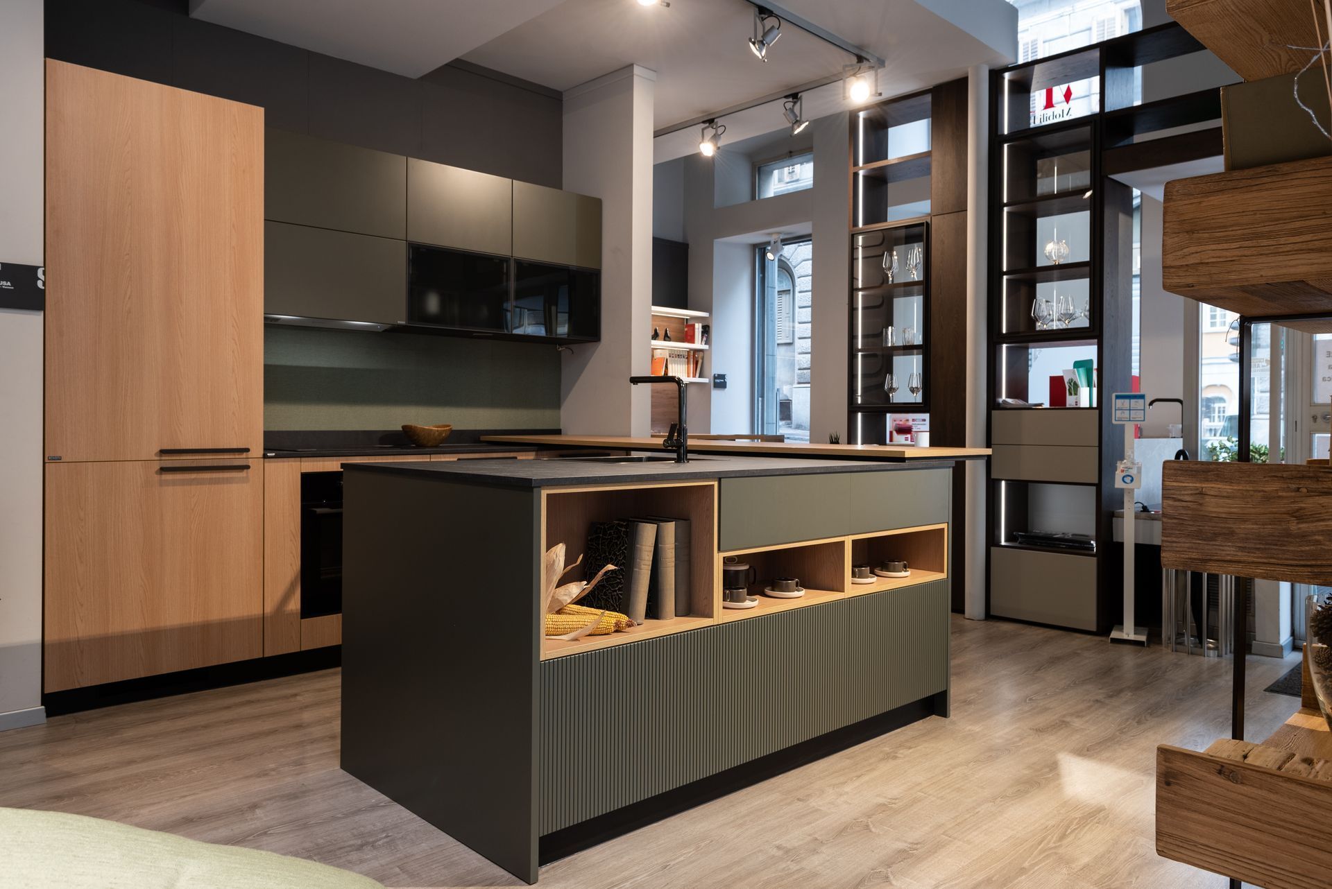 Cucina moderna con isola centrale, mobili scuri, dettagli in legno e ampie finestre che si affacciano su una stanza luminosa.