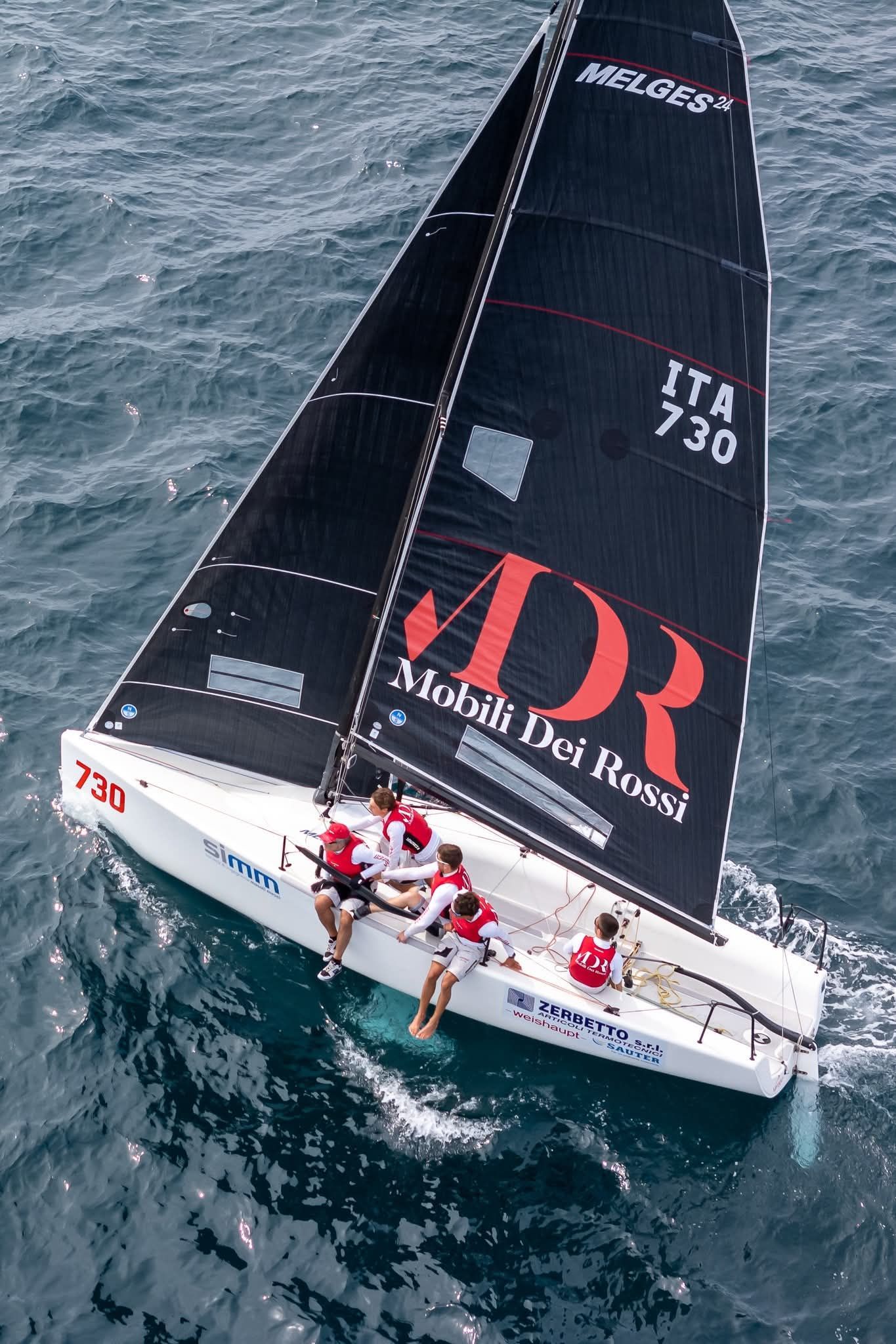 Barca a vela con vela nera e logo rosso che regata su acque agitate, equipaggio raggruppato a poppa.