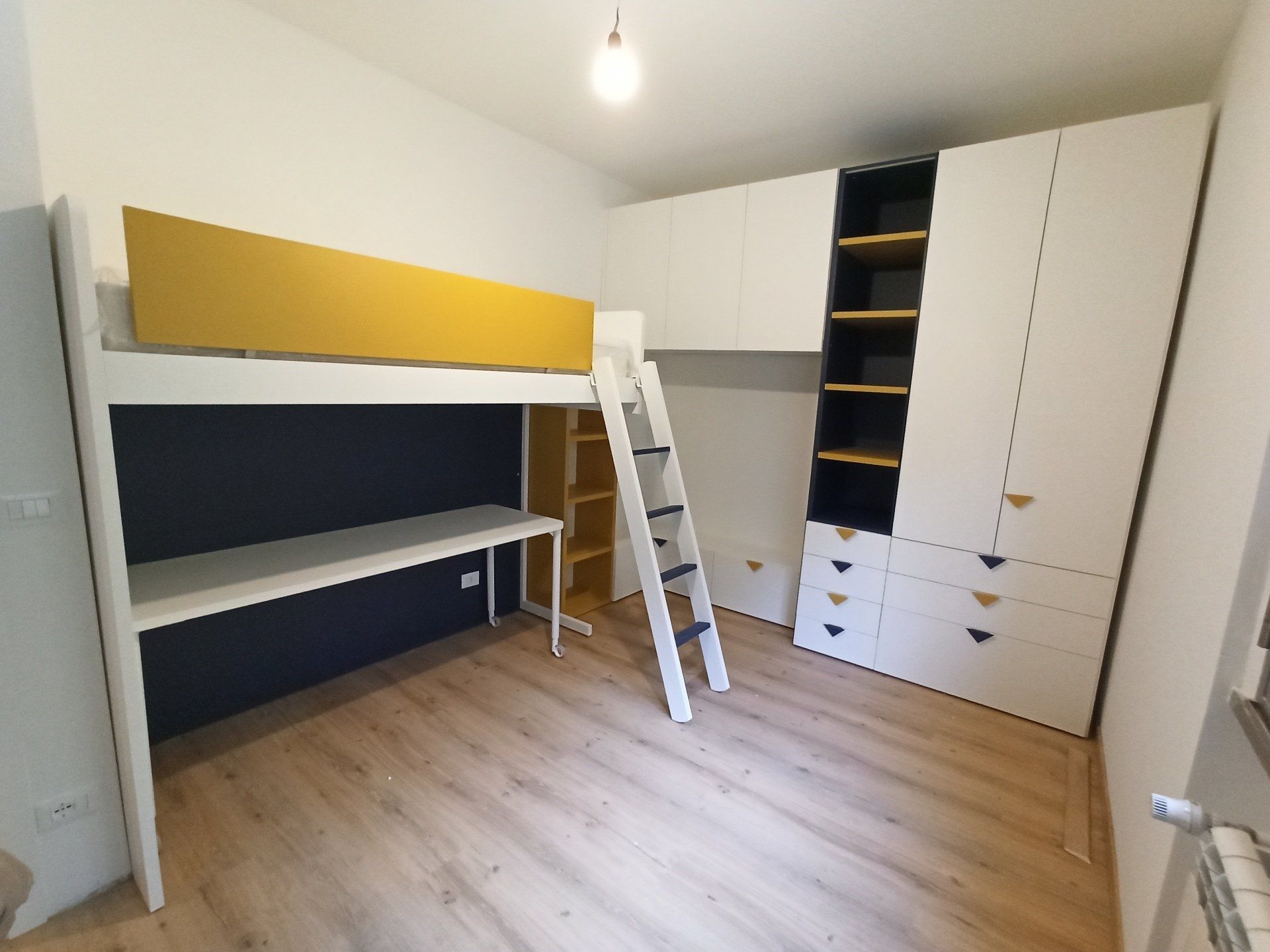 Piccola camera da letto con letto a soppalco, scrivania, scaletta e armadietti bianchi in una stanza luminosa