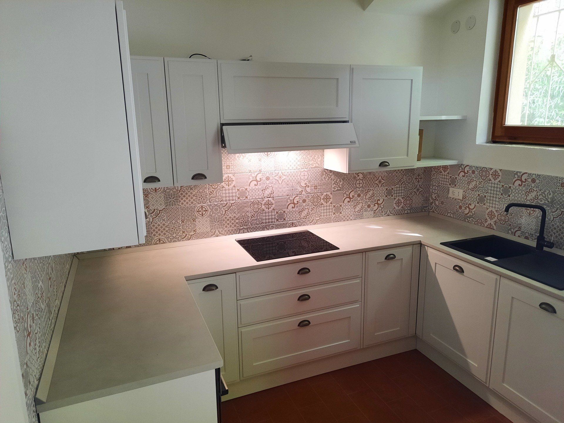 Cucina bianca con ripiani beige, paraschizzi piastrellato, piano cottura, cappa aspirante, lavello e mobili bianchi.