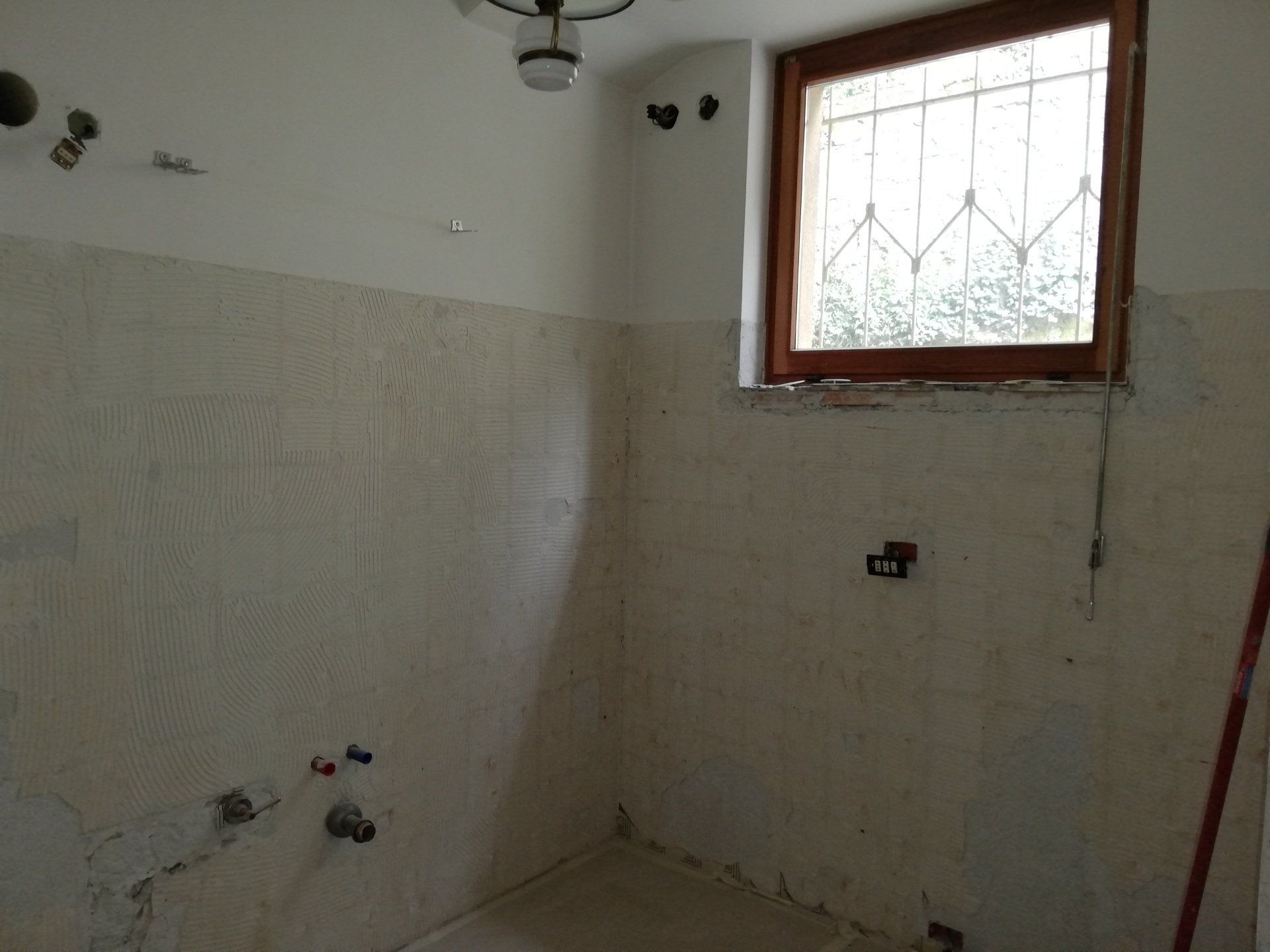 Bagno vuoto piastrellato in fase di ristrutturazione con tubature a vista e una piccola finestra