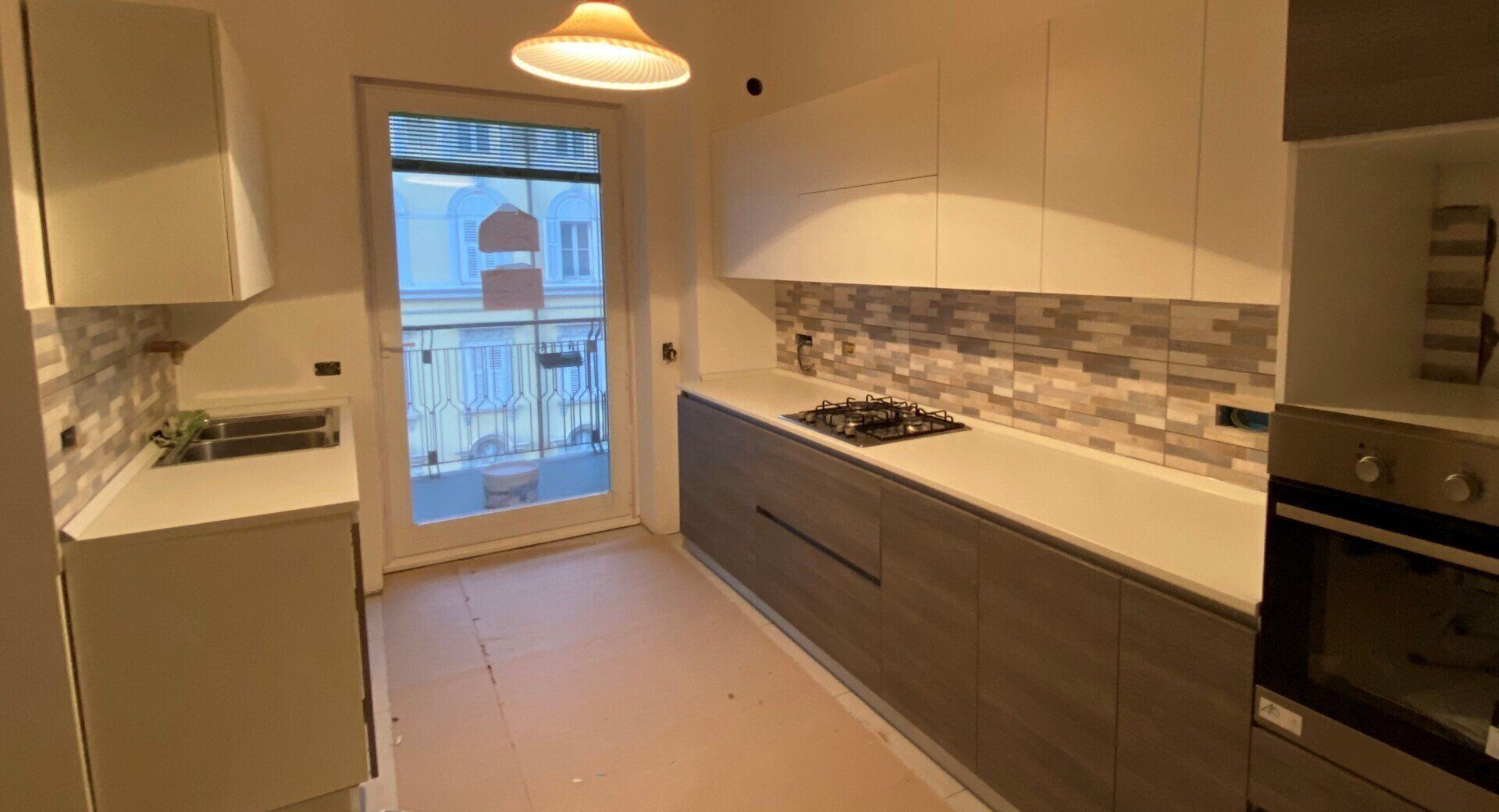 Cucina moderna e luminosa con mobili bianchi, cassetti inferiori scuri, paraschizzi piastrellato e porta che dà su un balcone.