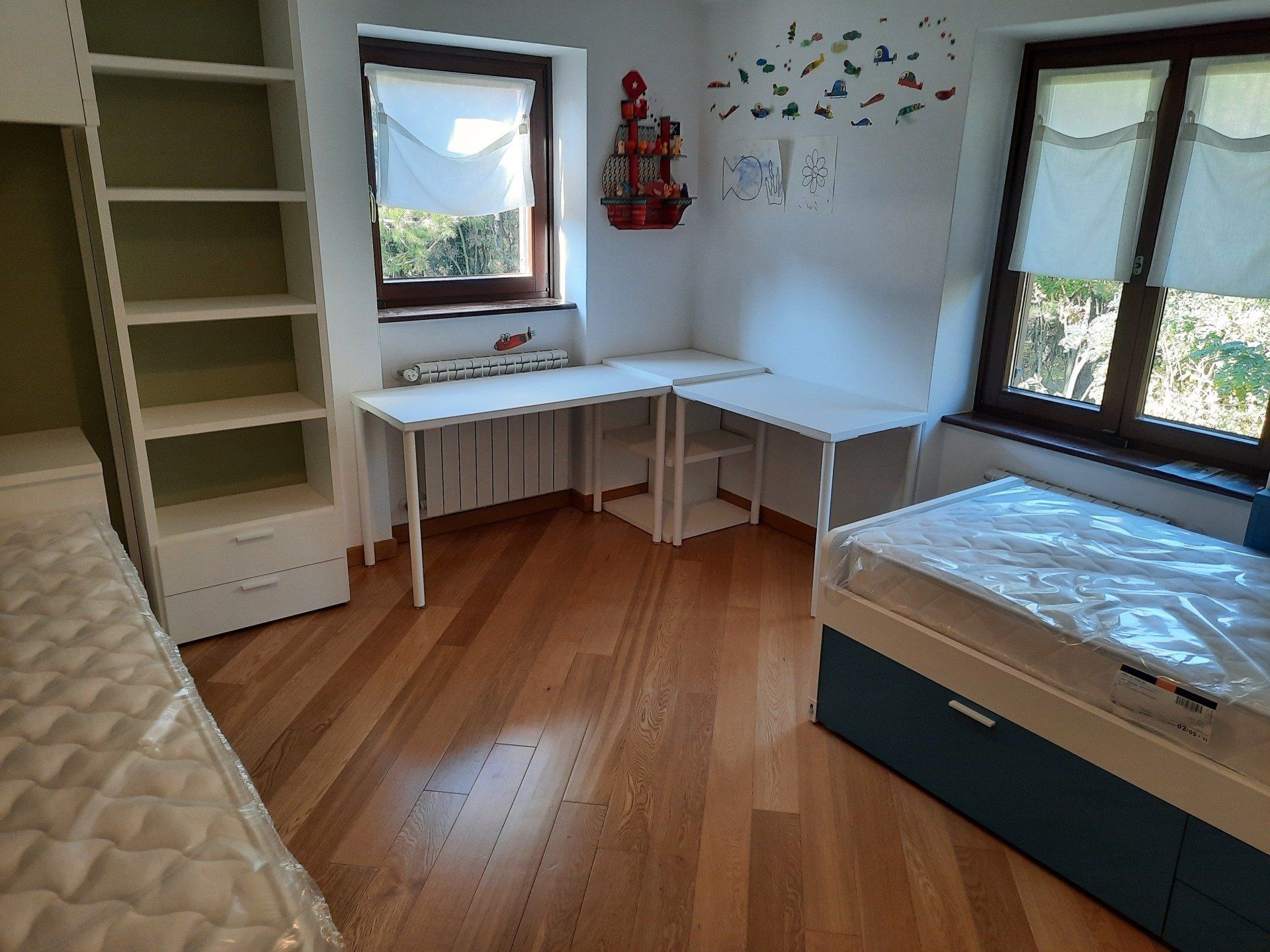 Piccola e luminosa camera da letto con scrivania, libreria, due finestre e un letto addossato alla parete.