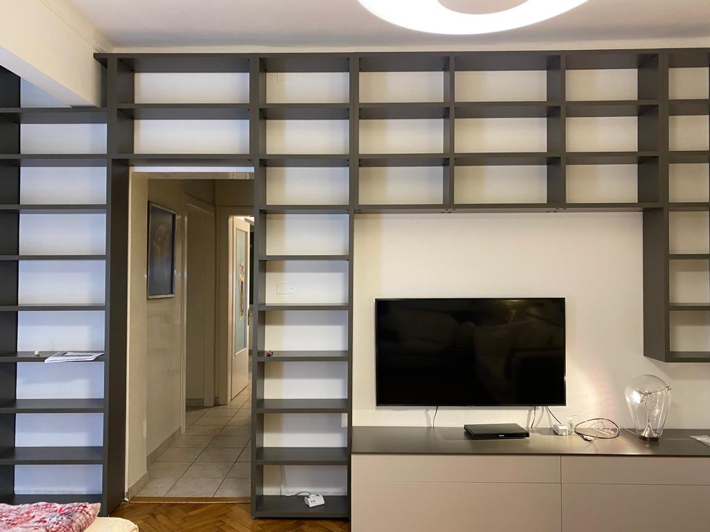 Soggiorno moderno con TV a parete, mensole grigie e corridoio visibile attraverso l'apertura centrale.