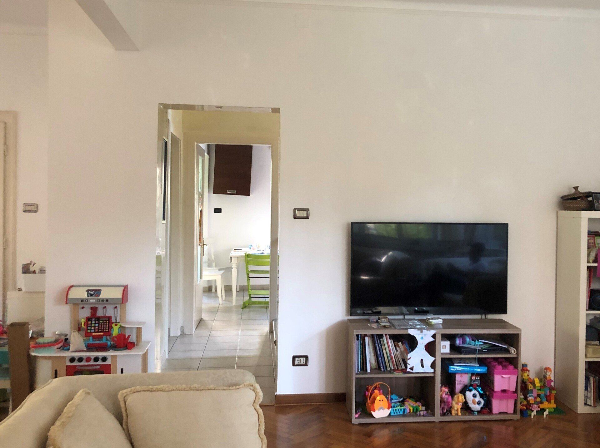 Soggiorno con divano beige, TV su un mobile, giocattoli e una porta che dà su un corridoio luminoso.
