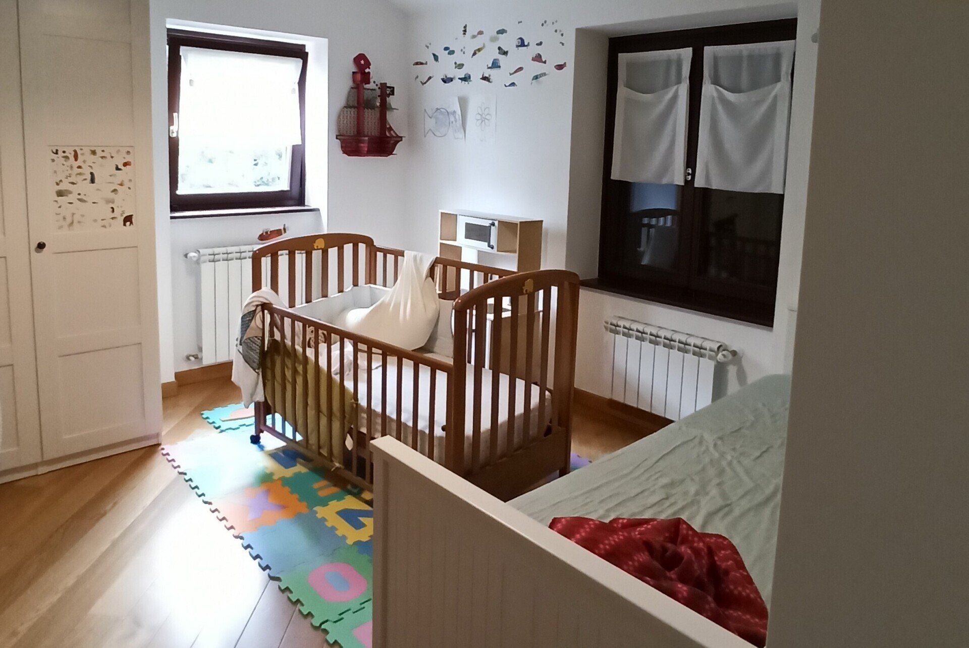 Cameretta luminosa con due culle in legno, un materassino in gommapiuma colorato, pareti bianche e una finestra con tende.