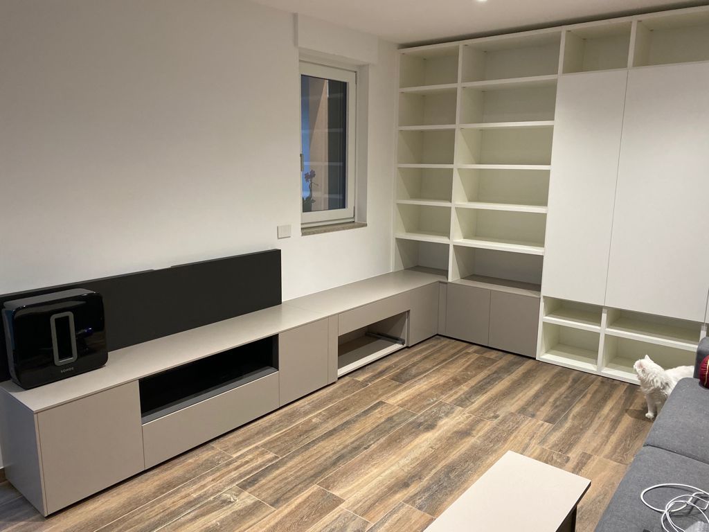 Soggiorno moderno con mensole bianche a muro, panca contenitore grigia, pavimento in legno e finestra.