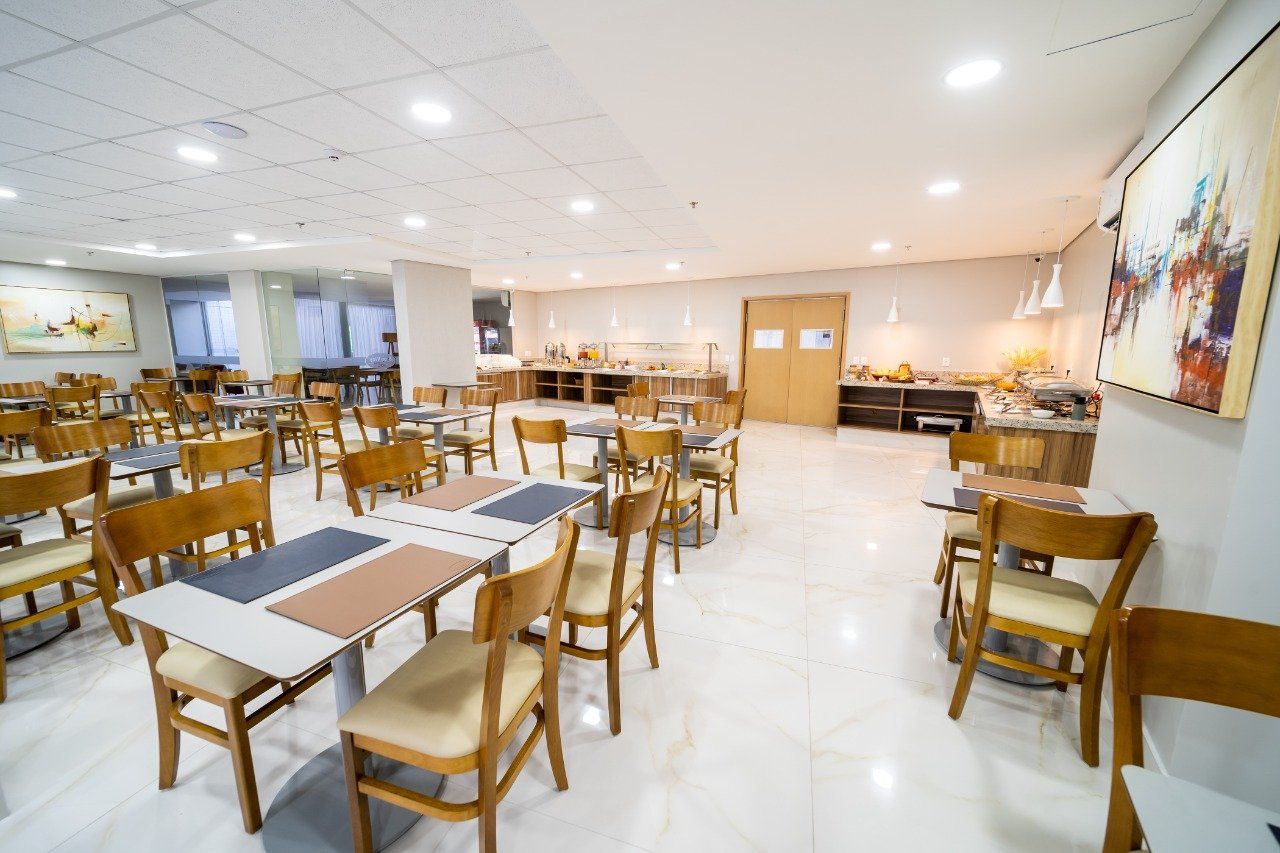 Sala de jantar vazia do hotel, com mesas, cadeiras e bufê de comida. Paredes e piso de cores claras.