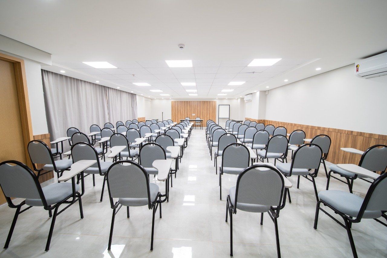 Uma sala de aula vazia com fileiras de cadeiras e carteiras, luzes brilhantes e painéis de madeira na parede.