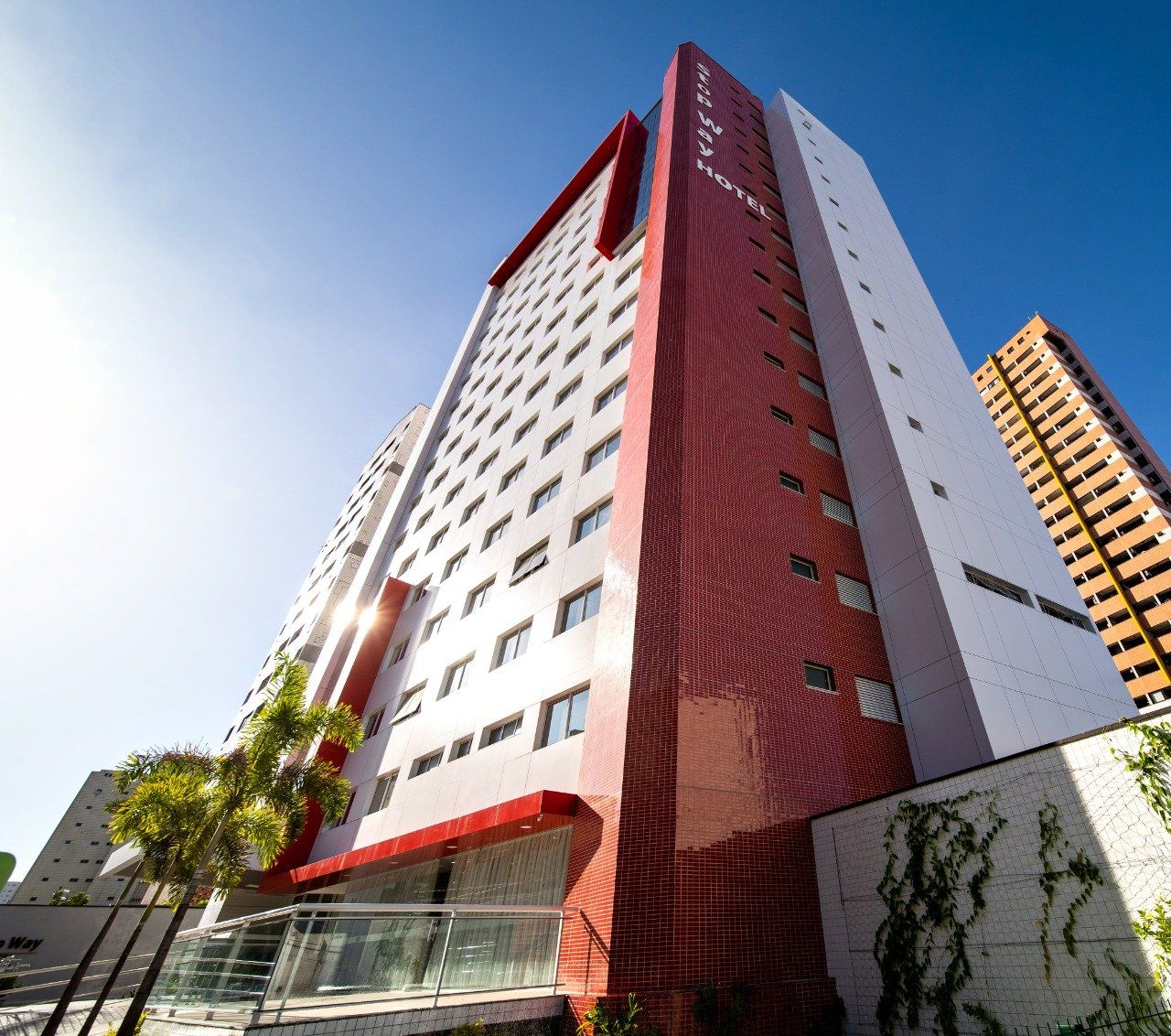 Exterior do hotel: edifício alto com detalhes em vermelho, fachada cinza e branca, palmeiras, céu azul claro.