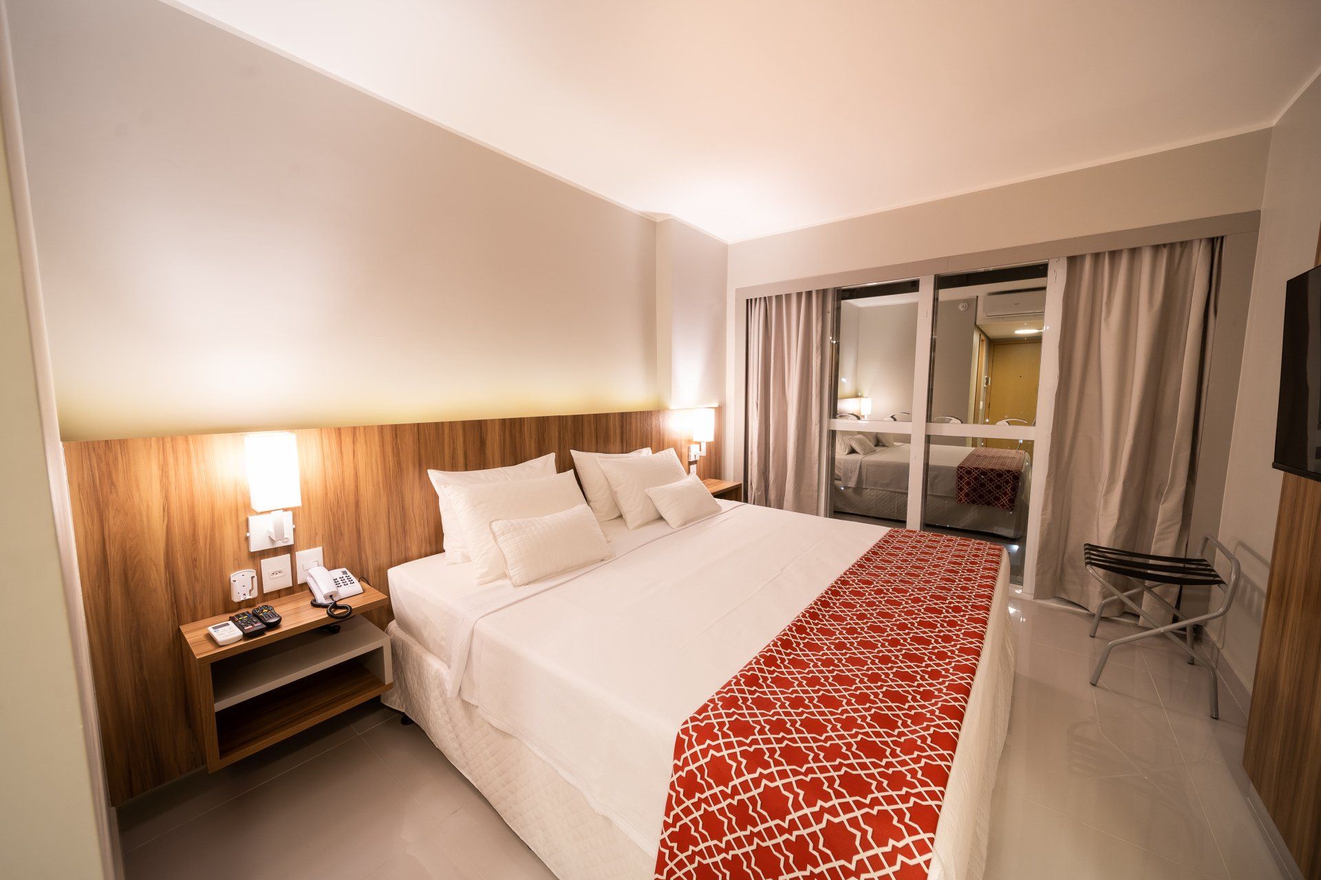 Quarto de hotel com cama king-size, tapete vermelho estampado, varanda e cores neutras.