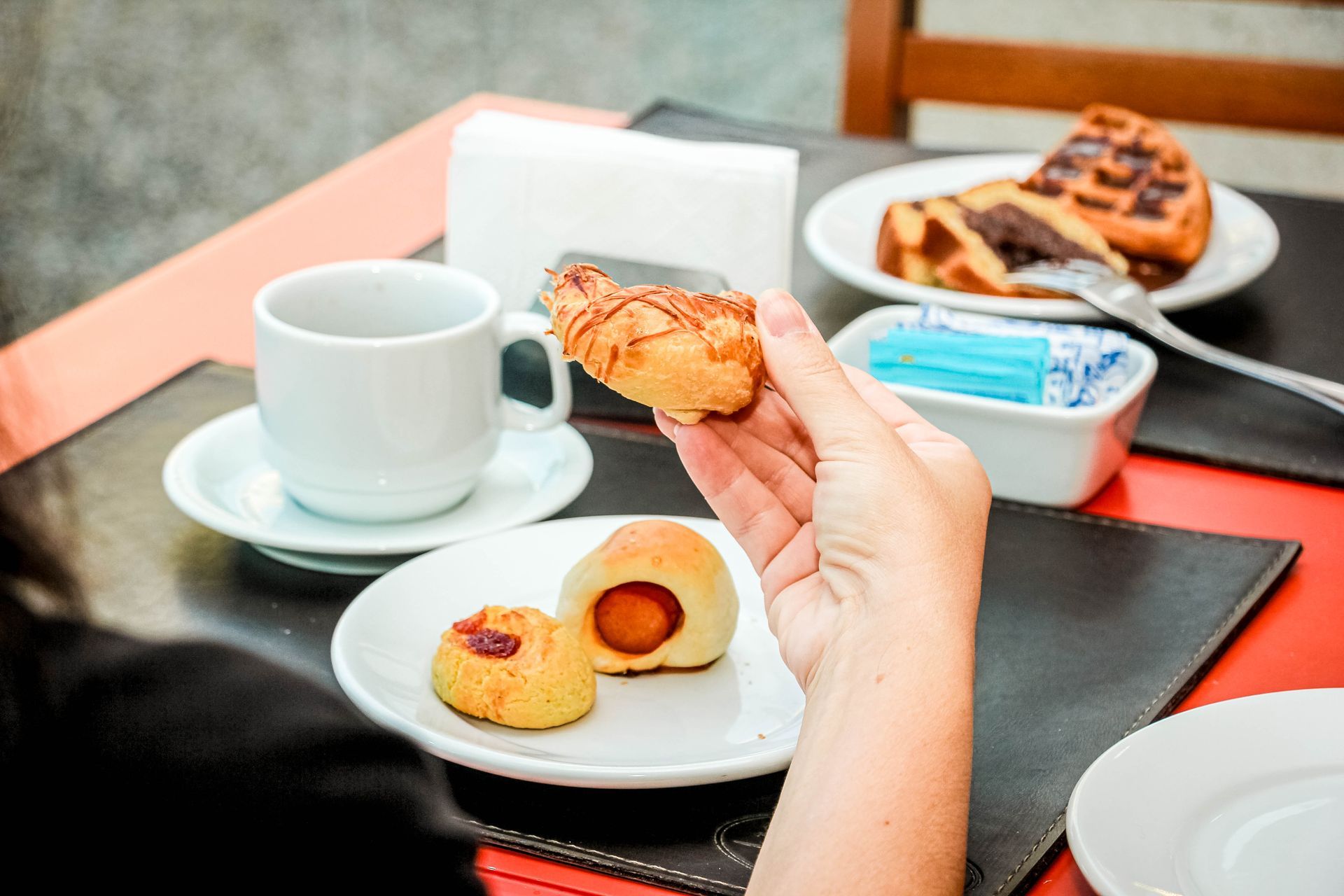 Mão de mulher segurando um pãozinho de salsicha empanado. Mesa de café da manhã com café, doces e waffles.