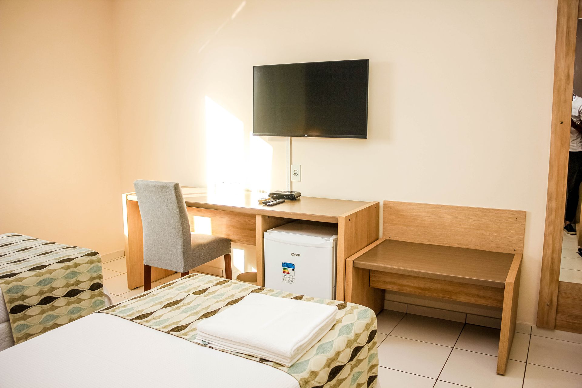 Quarto de hotel com camas, escrivaninha, TV, frigobar e um banco.
