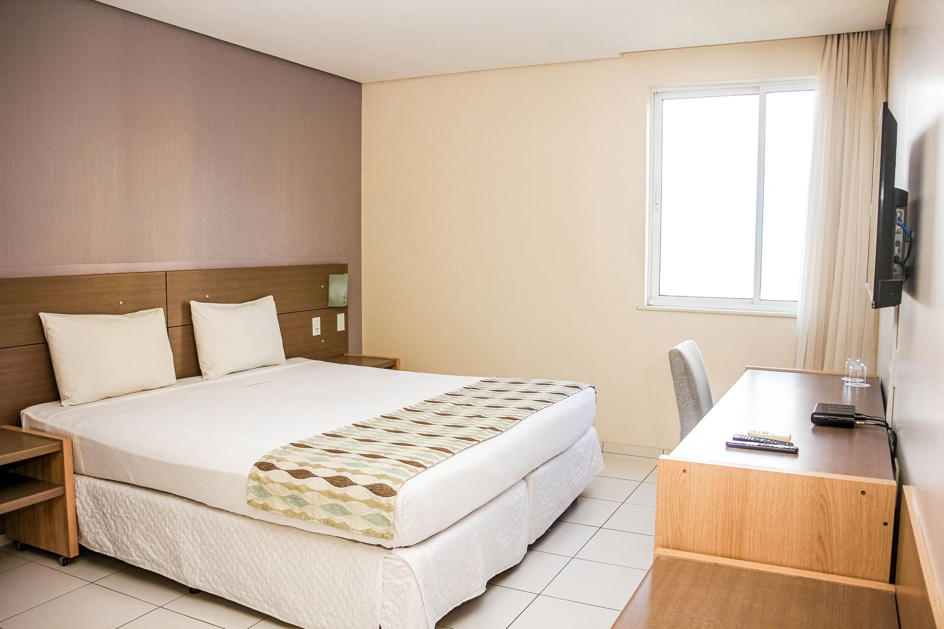 Quarto de hotel com cama, escrivaninha e janela.