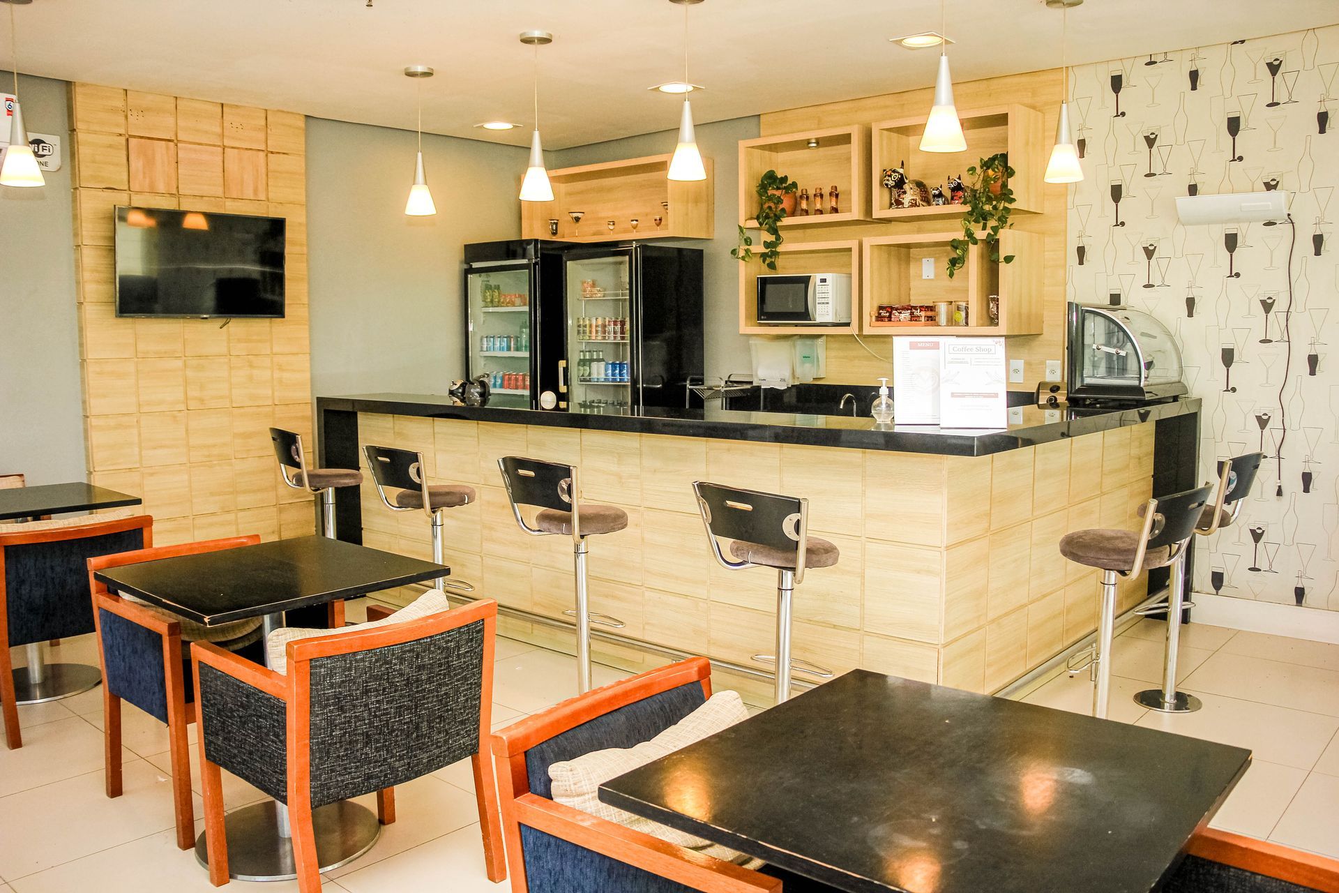 Um café com mesas, cadeiras, um bar, geladeiras e luzes penduradas.