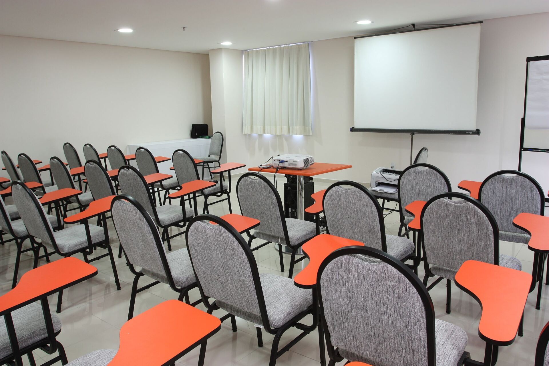 Sala de aula vazia com fileiras de cadeiras com braços de tablet laranja, tela de projeção e projetor.