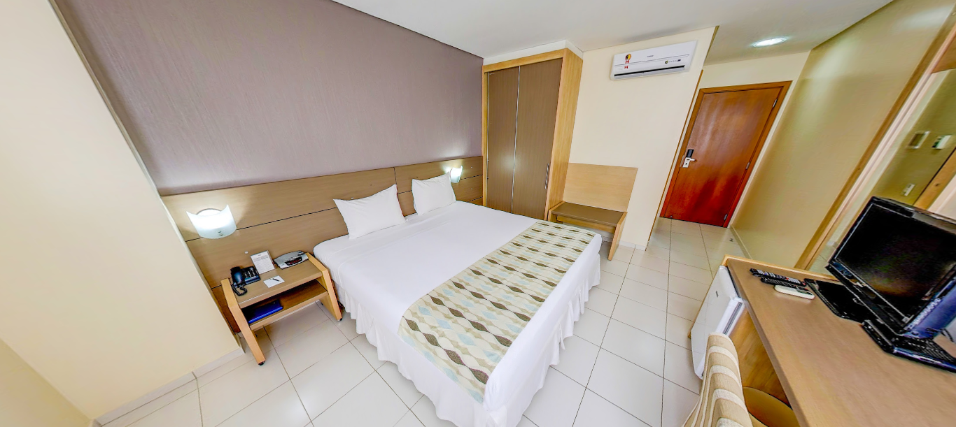 Quarto de hotel com cama, mesa de cabeceira, armário, escrivaninha e porta.
