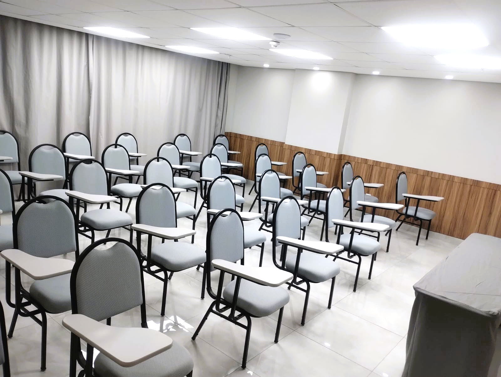 Sala de aula vazia com cadeiras cinzas, carteiras brancas e painéis de madeira.