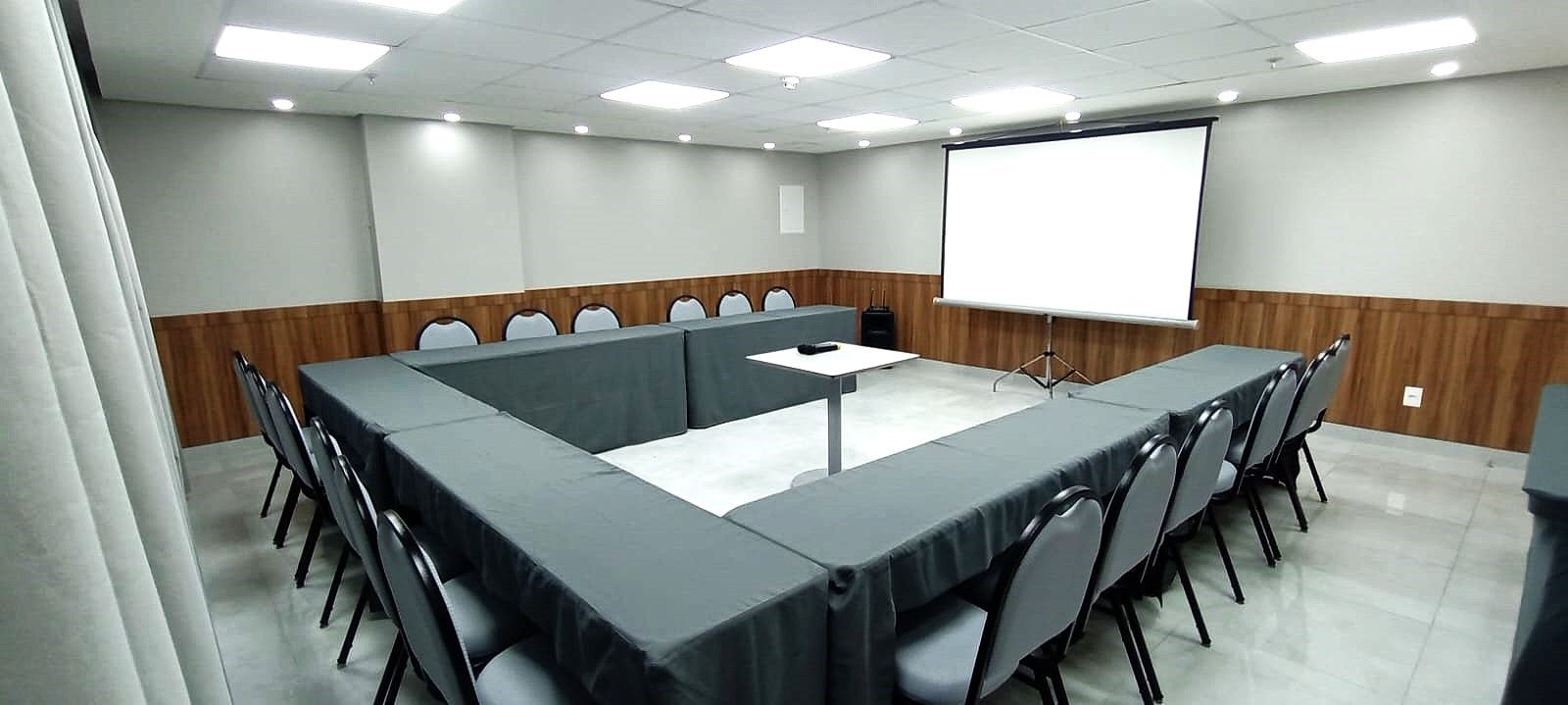 Sala de conferências com mesa em U, cadeiras e tela de projeção. Painéis de parede de madeira e esquema de cores neutras.