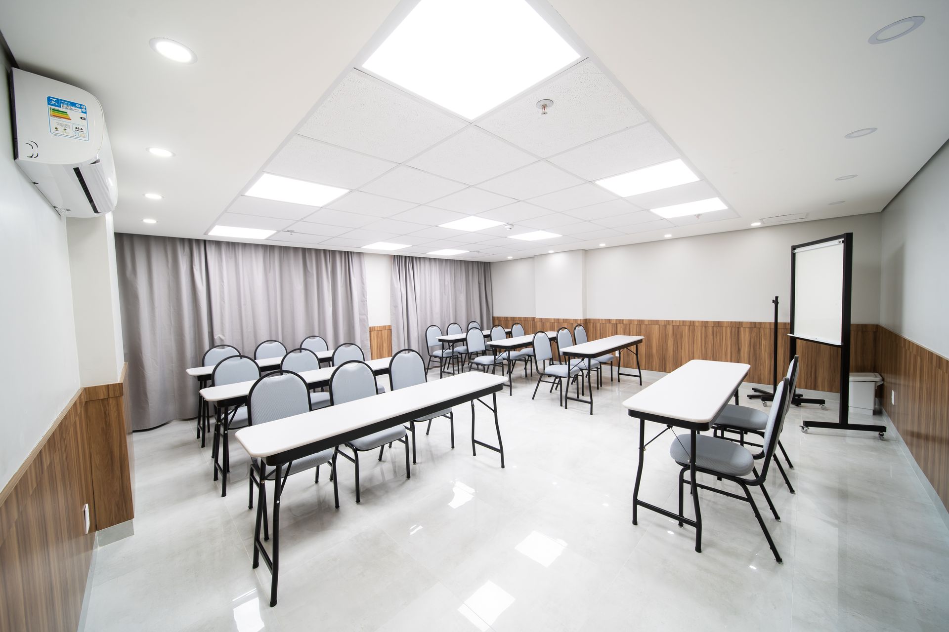 Sala de aula com cadeiras cinzas, mesas brancas e painéis de madeira nas paredes.