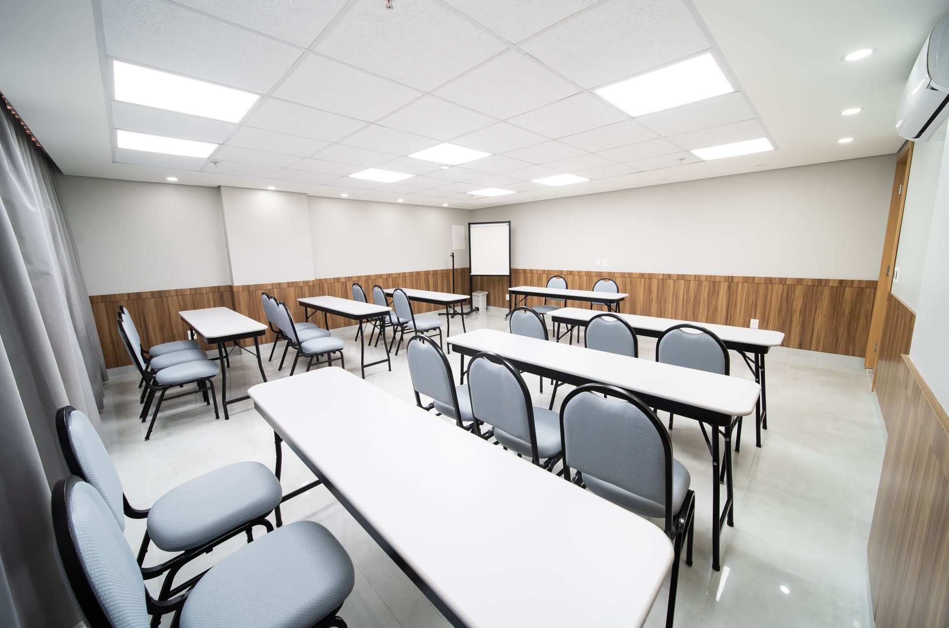 Sala de aula com mesas e cadeiras brancas, parede com painéis de madeira, quadro branco e iluminação suspensa.