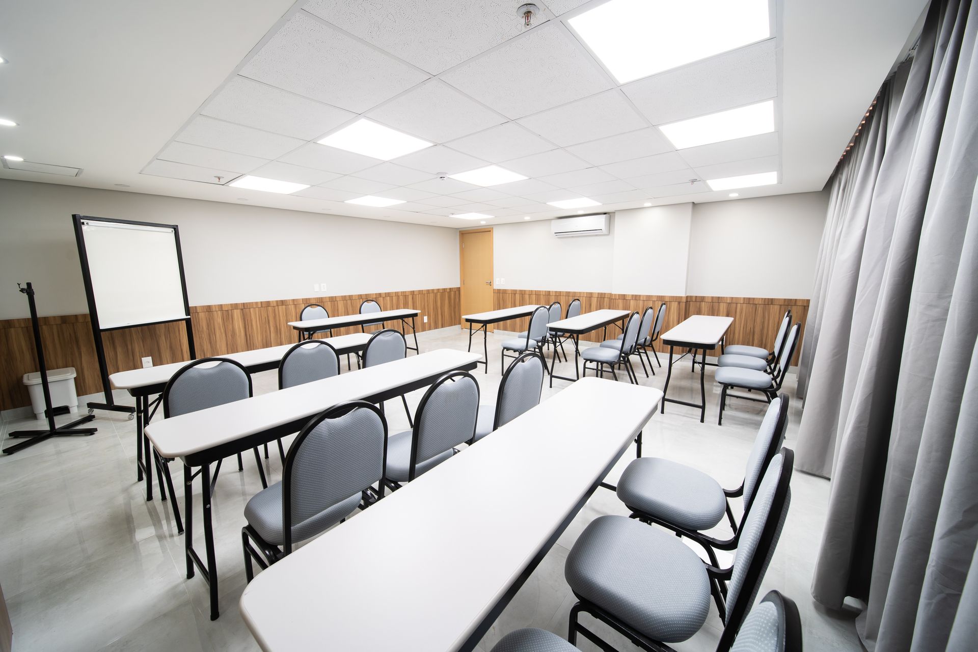 Sala de aula com mesas, cadeiras, quadro branco e cortinas. Teto branco, paredes com painéis de madeira.