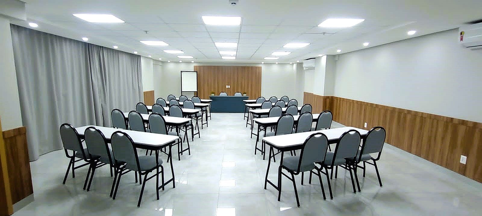 Sala de aula com fileiras de mesas e cadeiras voltadas para um palco. Paredes bege e brancas, cadeiras cinzas, luzes brilhantes no teto.