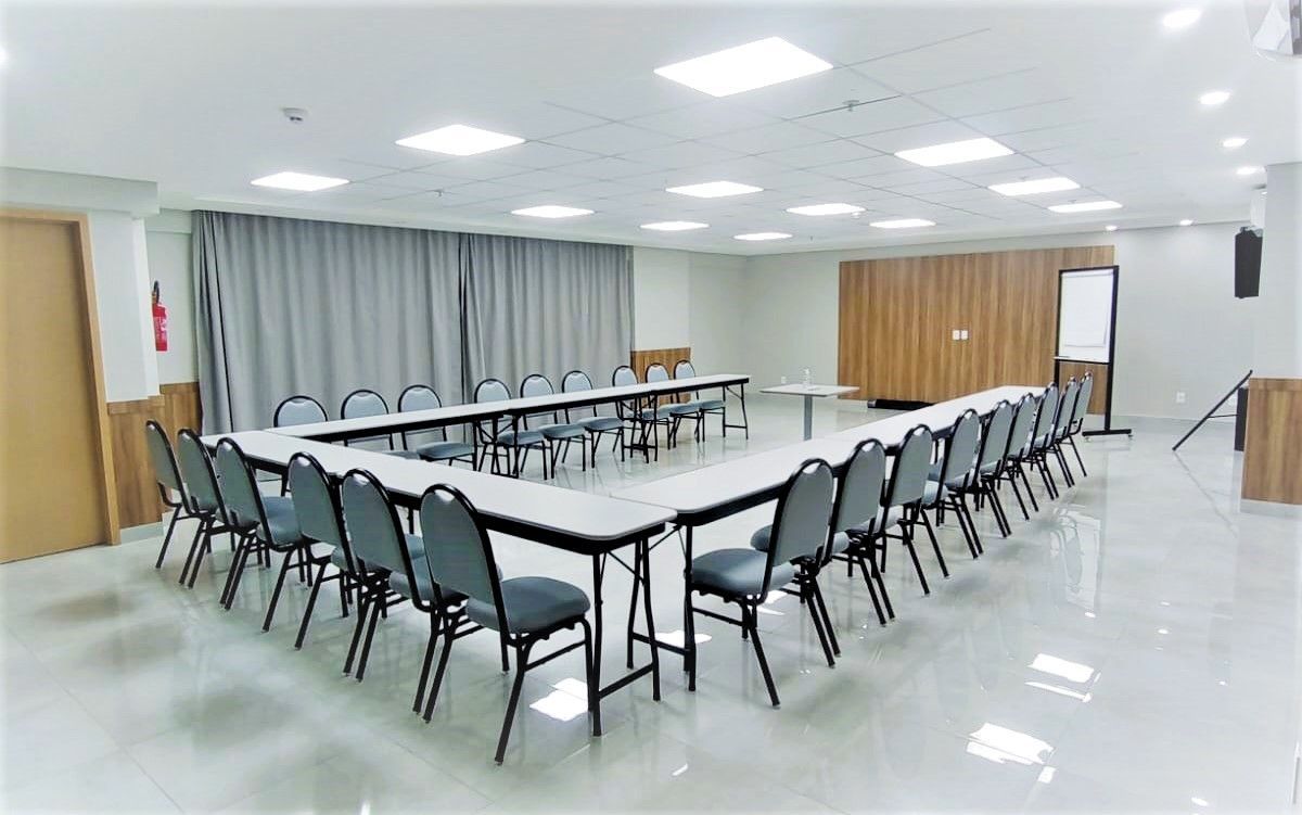 Sala de conferências com mesa e cadeiras em formato de U, cores neutras, piso branco e cortinas.