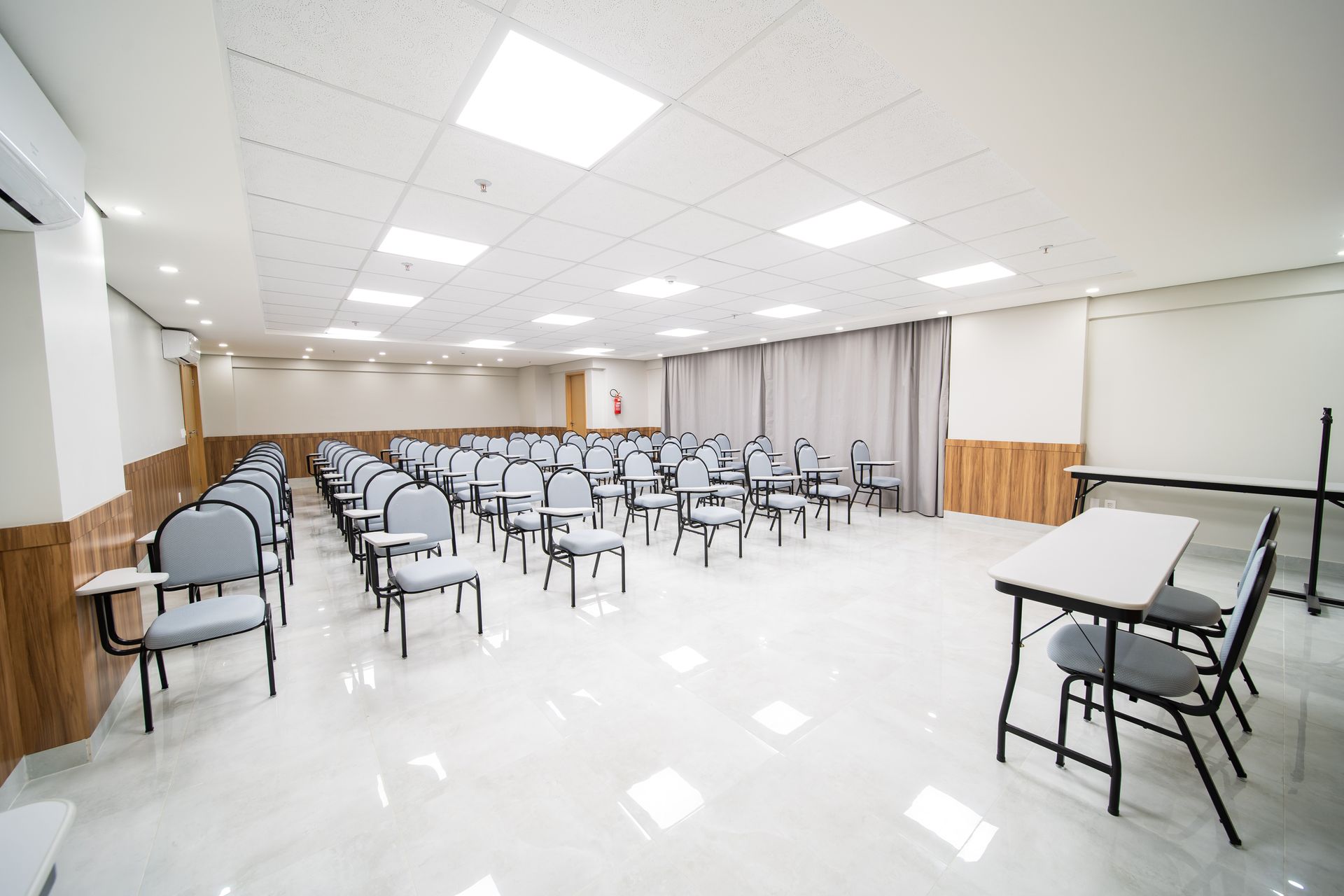 Uma sala de aula vazia com fileiras de cadeiras, uma mesa e uma tela de projetor. Teto e piso brancos brilhantes.